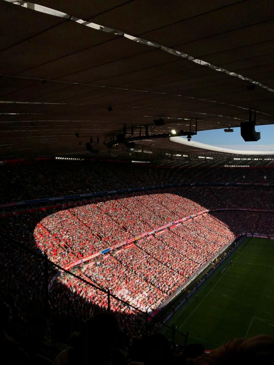 Vollbesetztes Stadion mit Fans in roten T-Shirts unter einem klaren Himmel, auf dem gr&uuml;nen Spielfeld im Vordergrund.