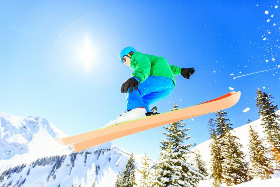 Ein Snowboarder springt über einen Schnee bedeckten Hang unter klarem blauen Himmel und strahlender Sonne.