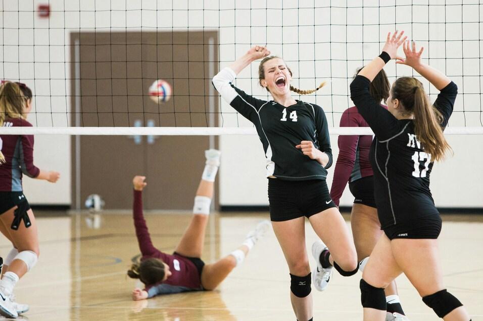 Zwei Volleyballspielerinnen feiern einen Punkt, w&auml;hrend eine andere Spielerin auf dem Boden liegt. Volleyballnetz im Hintergrund.