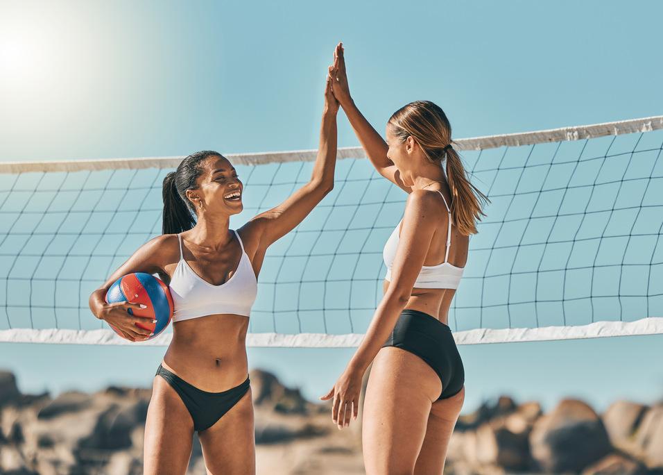 Zwei Frauen in Sportkleidung stehen am Volleyballnetz und feiern mit einem Ball in der Hand.