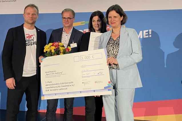 Vier Personen halten einen gro&szlig;en Scheck &uuml;ber 3.000 Euro, w&auml;hrend im Hintergrund ein Bildschirm den dritten Platz f&uuml;r &bdquo;Aktionsb&uuml;ndnis&ldquo; zeigt.