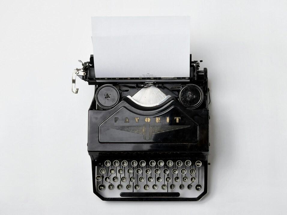 Schwarz-wei&szlig;e Schreibmaschine mit einem Blatt Papier, das aus der Typenhebeb&uuml;hne ragt, auf grauem Hintergrund.