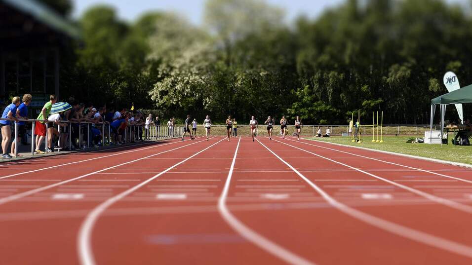 Athleten sprinten auf einer roten Laufbahn; Zuschauer beobachten vom Rand, Bäume im Hintergrund.