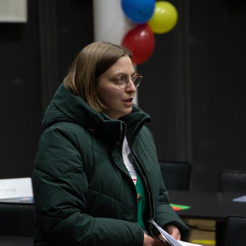 Frau spricht mit Mikrofon, tr&auml;gt eine gr&uuml;ne Jacke, vor bunten Luftballons in einem Konferenzraum.