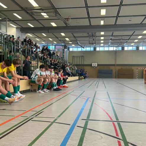 Jugendliche in Sportkleidung sitzen auf einer Bank in einer Sporthalle, Zuschauer und Trainer im Hintergrund.