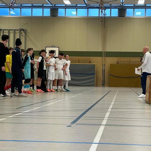 Gruppenspiele in einer Sporthalle: Jugendliche stehen in Uniformen, ein Trainer spricht zu ihnen, B&auml;lle liegen am Boden.