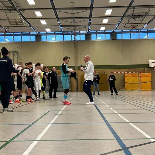 Jugendliche Sportsch&uuml;ler stehen in einer Halle, w&auml;hrend ein Trainer einem Spieler eine Auszeichnung &uuml;berreicht.