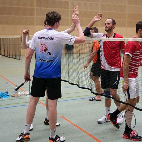 Gruppendynamik beim Badminton: Spieler geben sich nach einem Match High-Fives &uuml;ber dem Netz in einer Sporthalle.