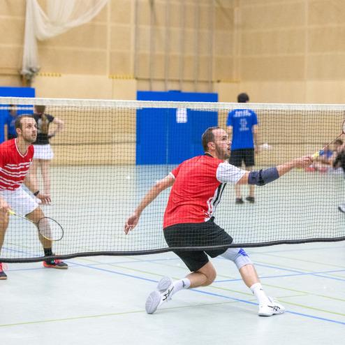 Zwei Badmintonspieler in roten Trikots w&auml;hrend eines Spiels auf einem Innenplatz, mit Netz und weiteren Spielern im Hintergrund.