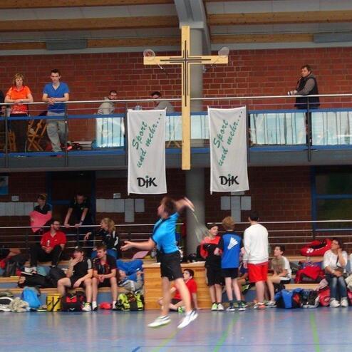Spieler in blauer Sportkleidung beim Badminton-Spiel in einer Halle mit Zuschauern und Bannern im Hintergrund.