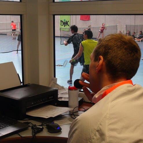 Blick auf eine Sporthalle mit Spielern im Badminton, w&auml;hrend ein Schiedsrichter am Tisch mit Mikrofon sitzt.