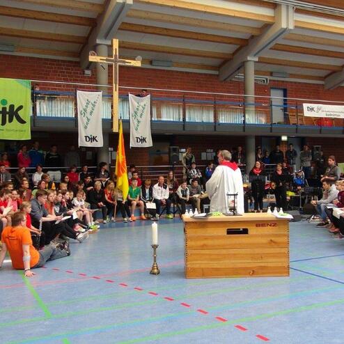 Gottesdienst in einer Sporthalle mit versammelten Jugendlichen, einem Priester und einem Altar mit Kerzen und Kreuz.