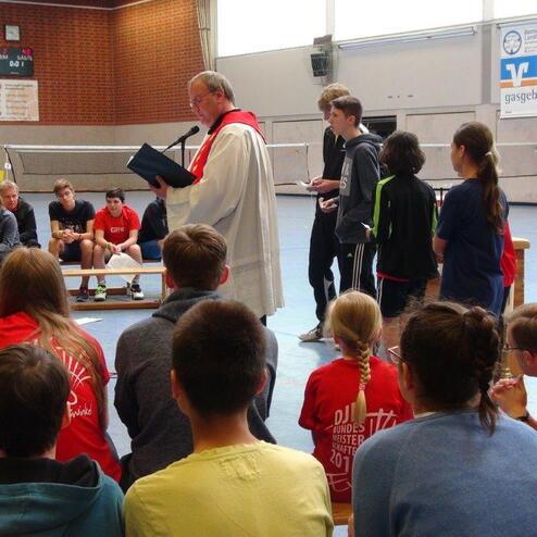 Ein Priester h&auml;lt eine Ansprache vor einer Gruppe von Jugendlichen in Sportkleidung in einer Turnhalle.