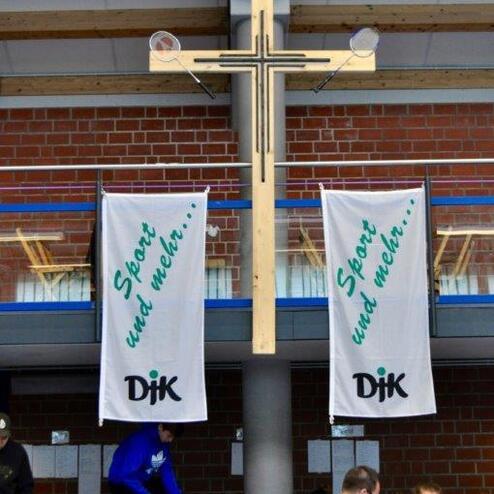 Holzkreuz &uuml;ber zwei Bannern mit der Aufschrift &bdquo;Sport und mehr&hellip;&ldquo; in einer Sporthalle, umgeben von Menschen.