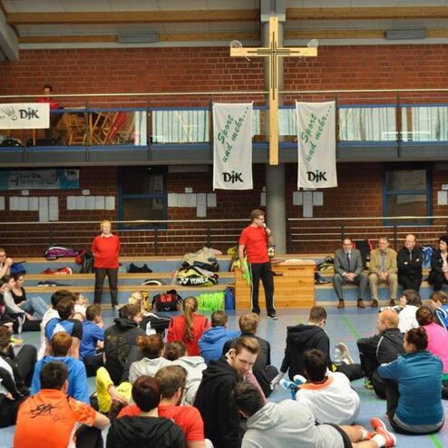 Versammlung in einer Sporthalle mit jungen Sportlern, die einem Redner in rotem Shirt zuh&ouml;ren, auf St&uuml;hlen sitzend.