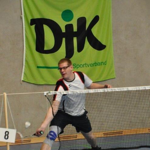 Spieler in Aktion beim Badminton, trifft den Ball mit dem Schl&auml;ger vor einem gro&szlig;en gr&uuml;nen DJK-Banner.