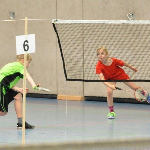 Zwei Kinder spielen Badminton in einer Sporthalle, eines schl&auml;gt den Federball &uuml;ber das Netz.
