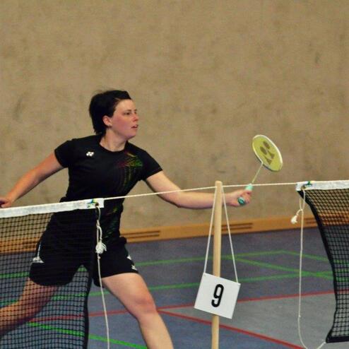 Frau schl&auml;gt einen Federball mit einem Schl&auml;ger in einem Sportraum, Netz und Punktetafel im Hintergrund sichtbar.