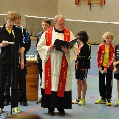 Gruppe von Kindern und Betreuern lauscht einem Redner in einer Sporthalle bei einer feierlichen Veranstaltung.