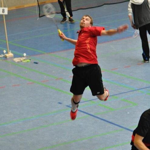 Spieler springt in rot-schwarzer Sportbekleidung mit einem Badminton-Schl&auml;ger in der Hand auf einem Badmintonfeld.