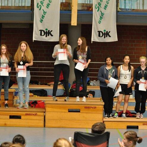 Gruppenaufnahme von acht M&auml;dchen auf einer B&uuml;hne, die Urkunden halten, vor einer Sporthalle mit Fahnen im Hintergrund.