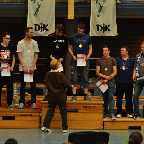 F&uuml;nf M&auml;nner auf Siegertreppen halten Urkunden, w&auml;hrend eine Frau ihnen Medaillen &uuml;berreicht. Sportveranstaltung in einer Halle.