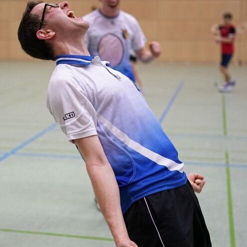 Ein Spieler jubelt in einem Sportoutfit, w&auml;hrend im Hintergrund weitere Spieler aktiv sind.