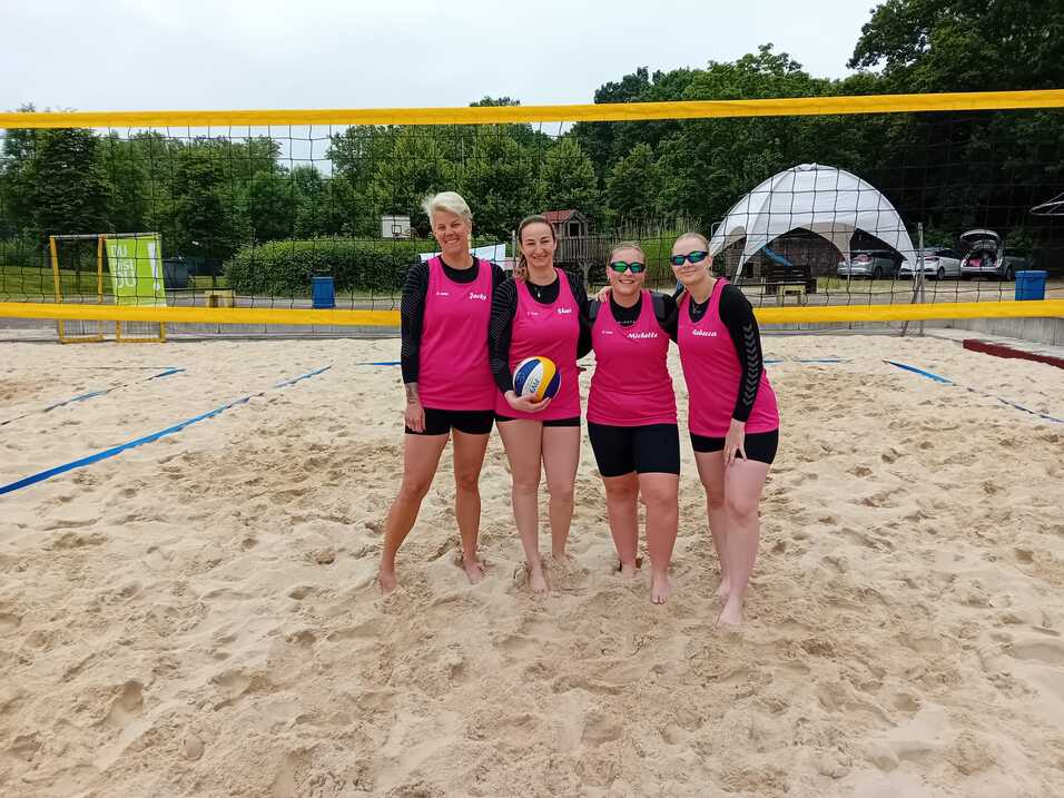 Vier Frauen in pinken Trikots stehen am Strand vor einem Volleyballnetz und halten einen Volleyball in der Hand.
