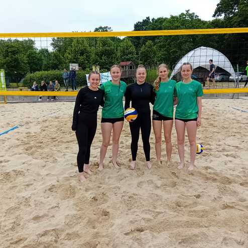 F&uuml;nf Volleyballspielerinnen in Trikots stehen l&auml;chelnd auf einem Sandplatz vor einem Volleyballnetz.