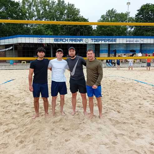 Vier M&auml;nner stehen im Sand der Beach Arena T&ouml;ppersee, umgeben von Volleyballnetzen und Zuschauern im Hintergrund.