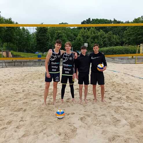 Vier Beachvolleyballspieler stehen auf einem Sandplatz, hinter ihnen ein Netz und gr&uuml;ne B&auml;ume im Hintergrund.