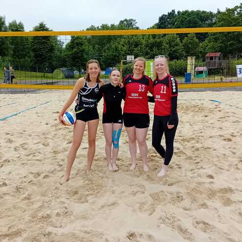 Vier Frauen in Sportbekleidung stehen auf einem Sandvolleyballfeld und halten einen Volleyball.