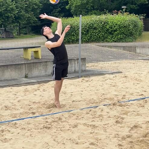 Junger Mann hebt beim Beachvolleyball einen Ball zum Aufschlag auf einem Sandplatz an.