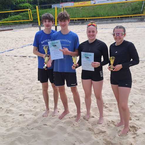 Vier Jugendliche stehen auf einem Sandplatz, halten Pokale und Urkunden nach einem Beachvolleyball-Turnier.
