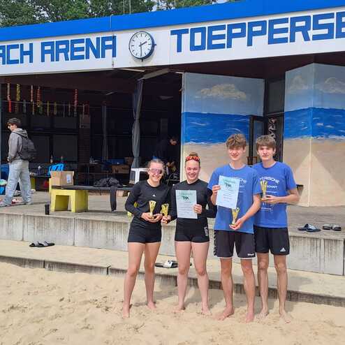 Vier Jugendliche halten Pokale und Urkunden vor der Beach Arena T&ouml;ppensee, mit Sand und Geb&auml;udewand im Hintergrund.