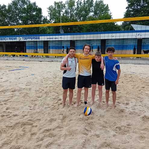 Vier Personen stehen am Strand vor einem Volleyballnetz, ein Ball liegt im Sand zwischen ihnen.