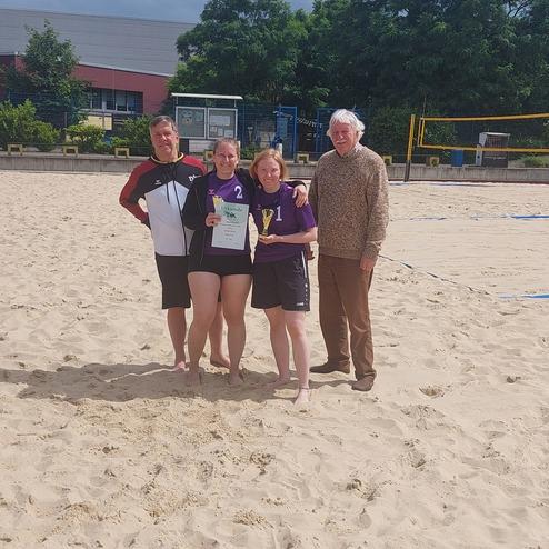 Vier Personen stehen auf einer Sandfl&auml;che, zwei Sportlerinnen halten ein Zertifikat, w&auml;hrend zwei M&auml;nner daneben l&auml;cheln.