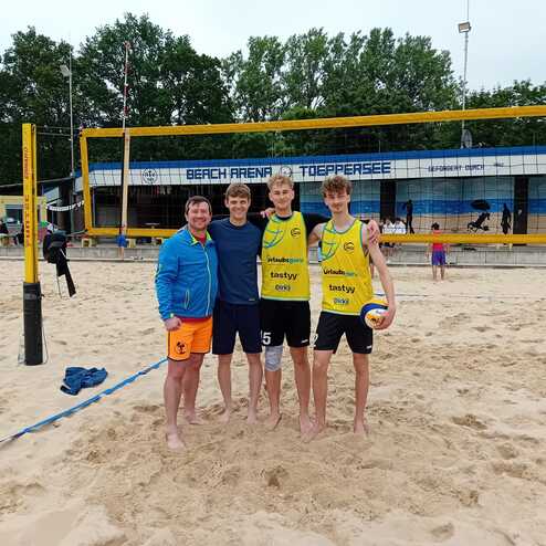 Vier Personen stehen l&auml;chelnd im Sand einer Beachvolleyball-Arena. Im Hintergrund sind Volleyballnetze und Zuschauer sichtbar.