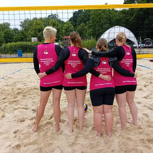Vier Frauen stehen im Sand hinter einem Volleyballnetz, alle tragen pinke Trikots und schauen in Richtung Spielfeld.