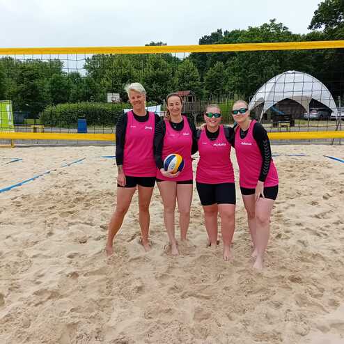 Vier Frauen in pinken Trikots stehen am Strand vor einem Volleyballnetz und halten einen Volleyball in der Hand.