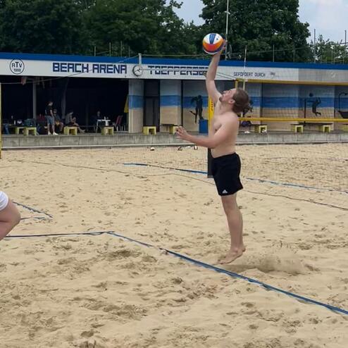 Spieler springt am Strand in der Beach Arena und schl&auml;gt einen Volleyball &uuml;ber das Netz. Zuschauer sind im Hintergrund sichtbar.