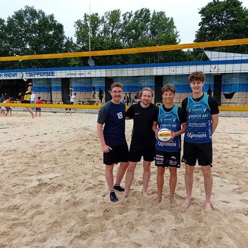 Vier Personen stehen l&auml;chelnd auf einem Sandstrand vor einem Beachvolleyballnetz.