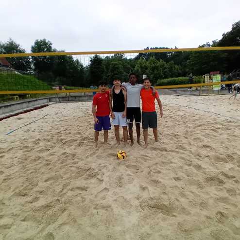 Vier Personen stehen auf einem Sandplatz f&uuml;r Beachvolleyball, mit netz und Ball im Hintergrund.