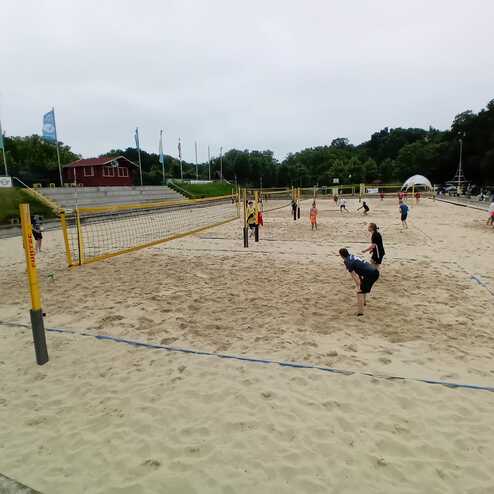 Mehrere Personen spielen Beachvolleyball auf einem Sandplatz bei bew&ouml;lktem Wetter. Im Hintergrund sind Fahnen zu sehen.