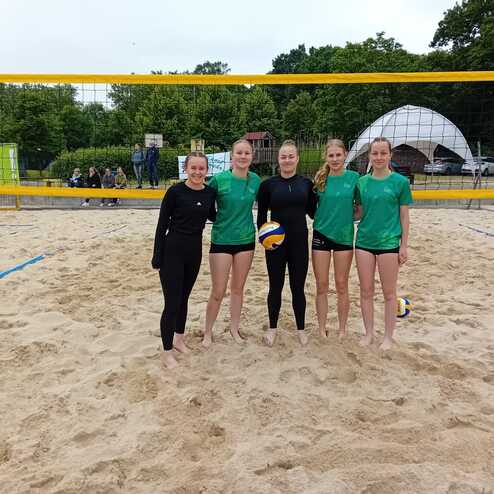 F&uuml;nf weibliche Volleyballspielerinnen in Sportkleidung stehen am Sandplatz hinter dem Netz und l&auml;cheln in die Kamera.