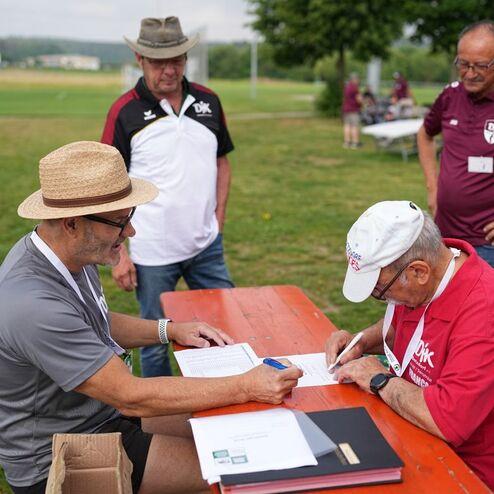 Zwei M&auml;nner sitzen an einem Tisch im Freien, w&auml;hrend zwei weitere im Hintergrund stehen und zuschauen.