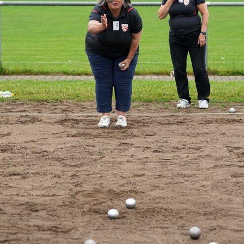 Frau wirft eine Kugel auf einem Bouleplatz, w&auml;hrend eine andere Frau zuschaut und mit der Hand gestikuliert.