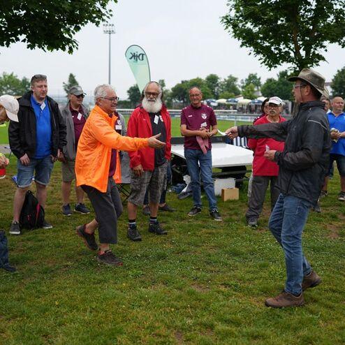 Gruppenszene mit mehreren Menschen auf einer Wiese, w&auml;hrend zwei Personen in Interaktion sind.