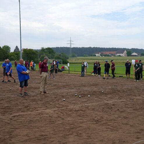 Gruppenspiel auf einem sandigen Spielfeld mit mehreren Personen, die Boule-Kugeln werfen, Zuschauer im Hintergrund.