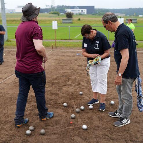 Drei Personen stehen auf einem Sandplatz und beurteilen die Platzierung von Boulekugeln.